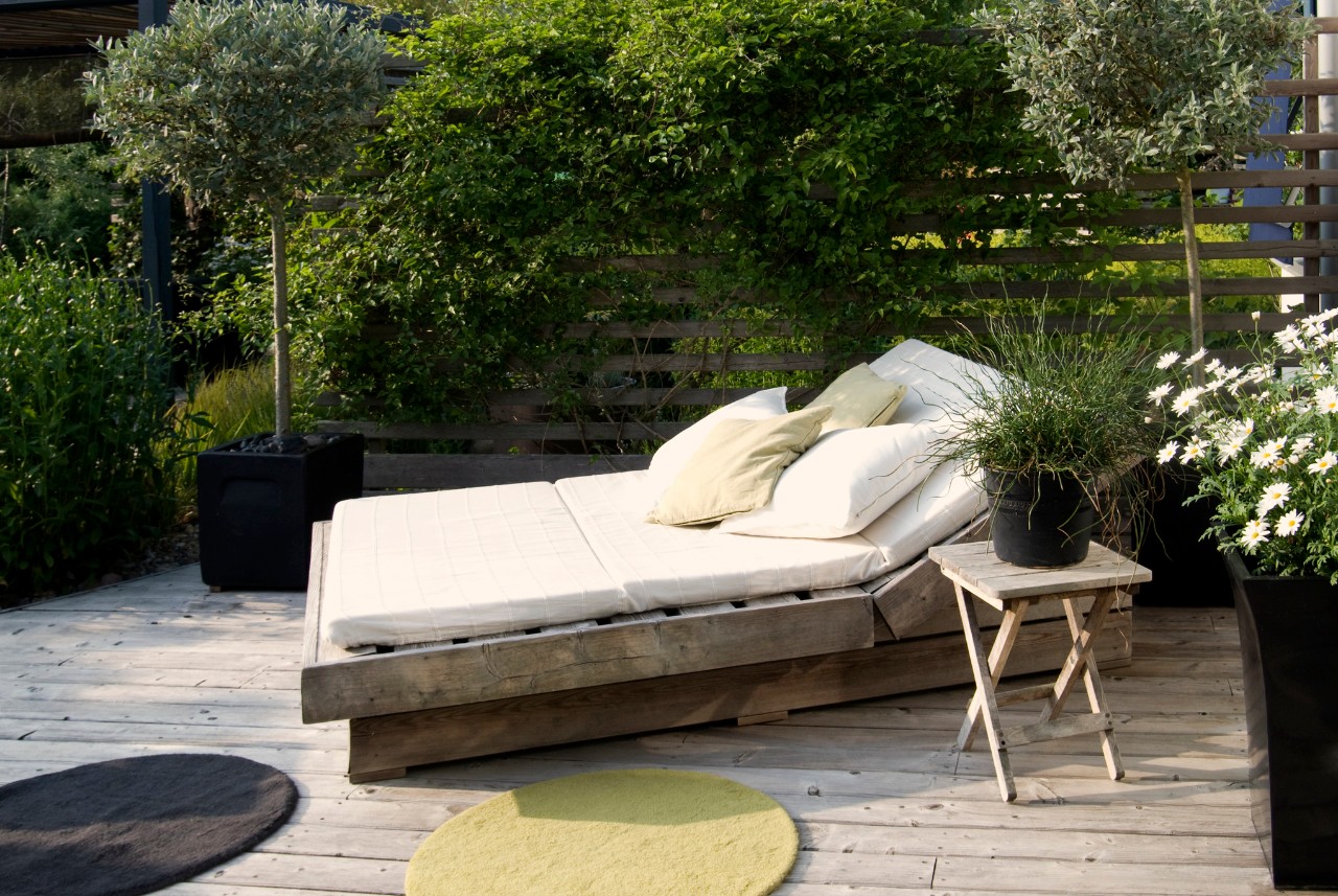 Une terrasse en bois avec une chaise longue et un matelas, des oliviers en arrière-plan