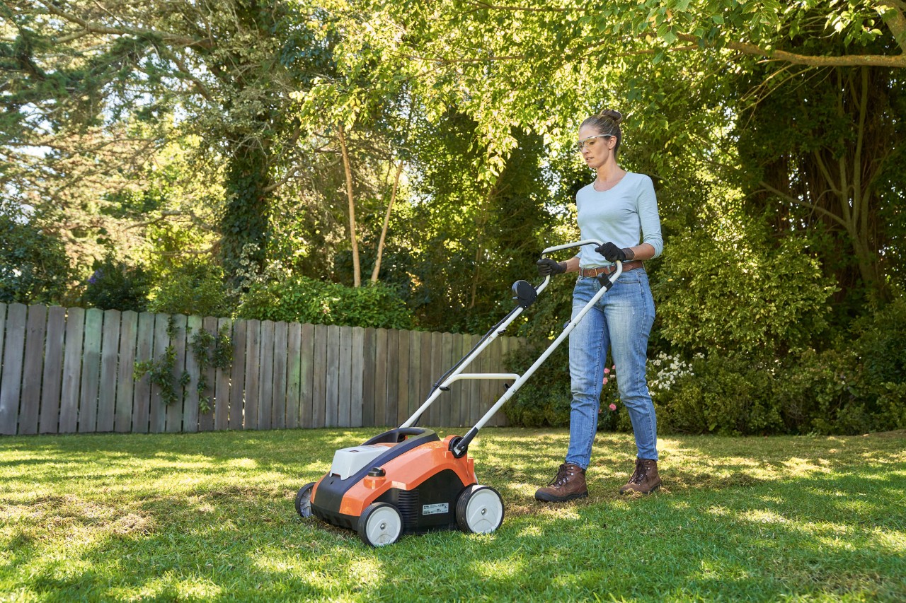 Une femme aère le gazon avec le scarificateur sur batterie STIHL RLA 240 dans un jardin, équipée de lunettes de protection et de gants de travail.