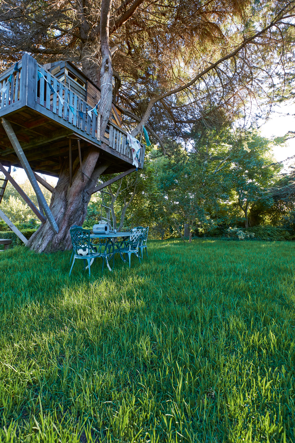 Un terrain verdoyant avec une cabane dans un arbre, une table et des chaises de jardin vertes et de grands arbres