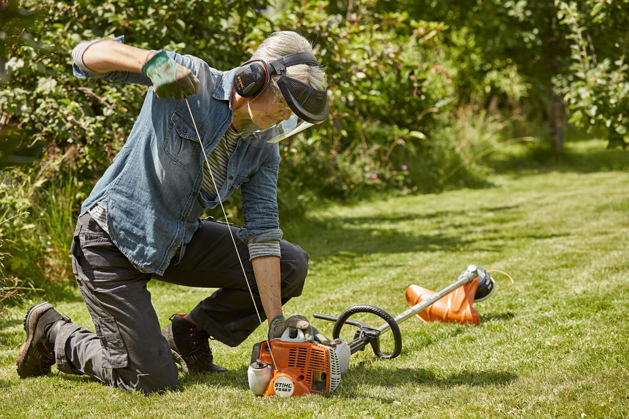 Une femme portant des protections auditive et faciale est en train de démarrer une débroussailleuse à essence STIHL FS&nbsp;56&nbsp;R sur un terrain vert