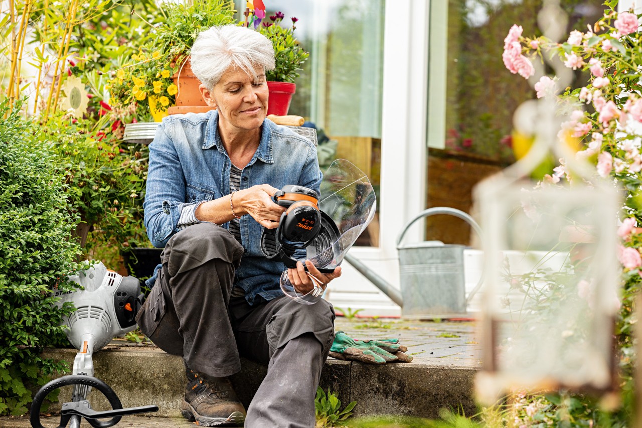 Une femme est assise entre des plantes en fleurs à côté d’une débroussailleuse STIHL FS&nbsp;38 et examine l’équipement de protection