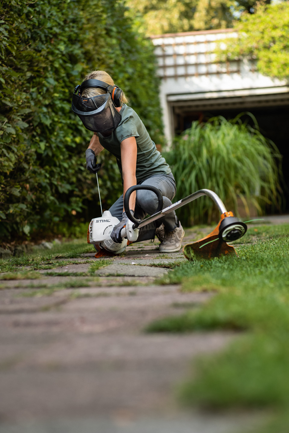 Une femme portant un équipement de protection coupe-bordure et des gants en train de démarrer un coupe-bordure thermique STIHL FS 38 posé sur un chemin dans un jardin
