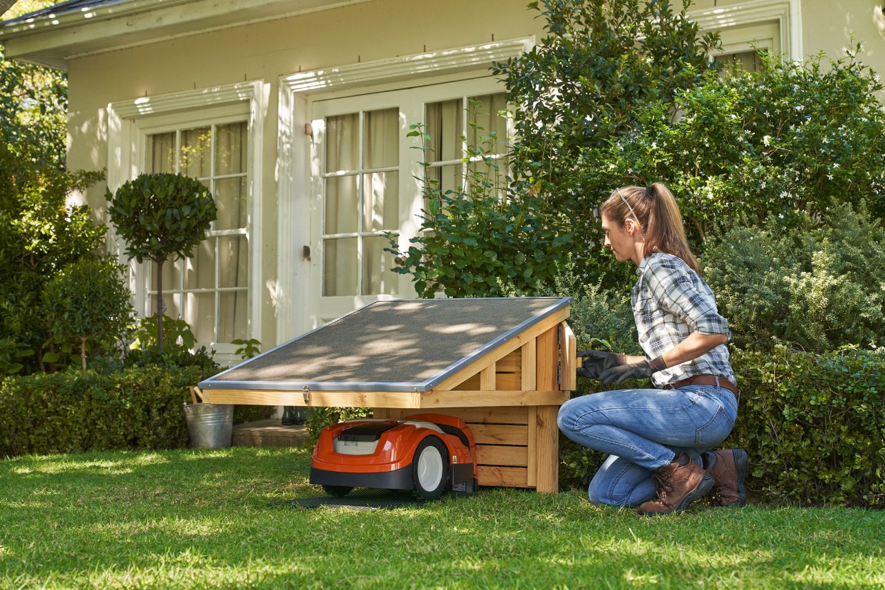 Une femme avec un équipement de protection s’agenouille à côté d’un abri STIHL iMOW® où se trouve un robot de tonte et ouvre le compartiment de rangement 