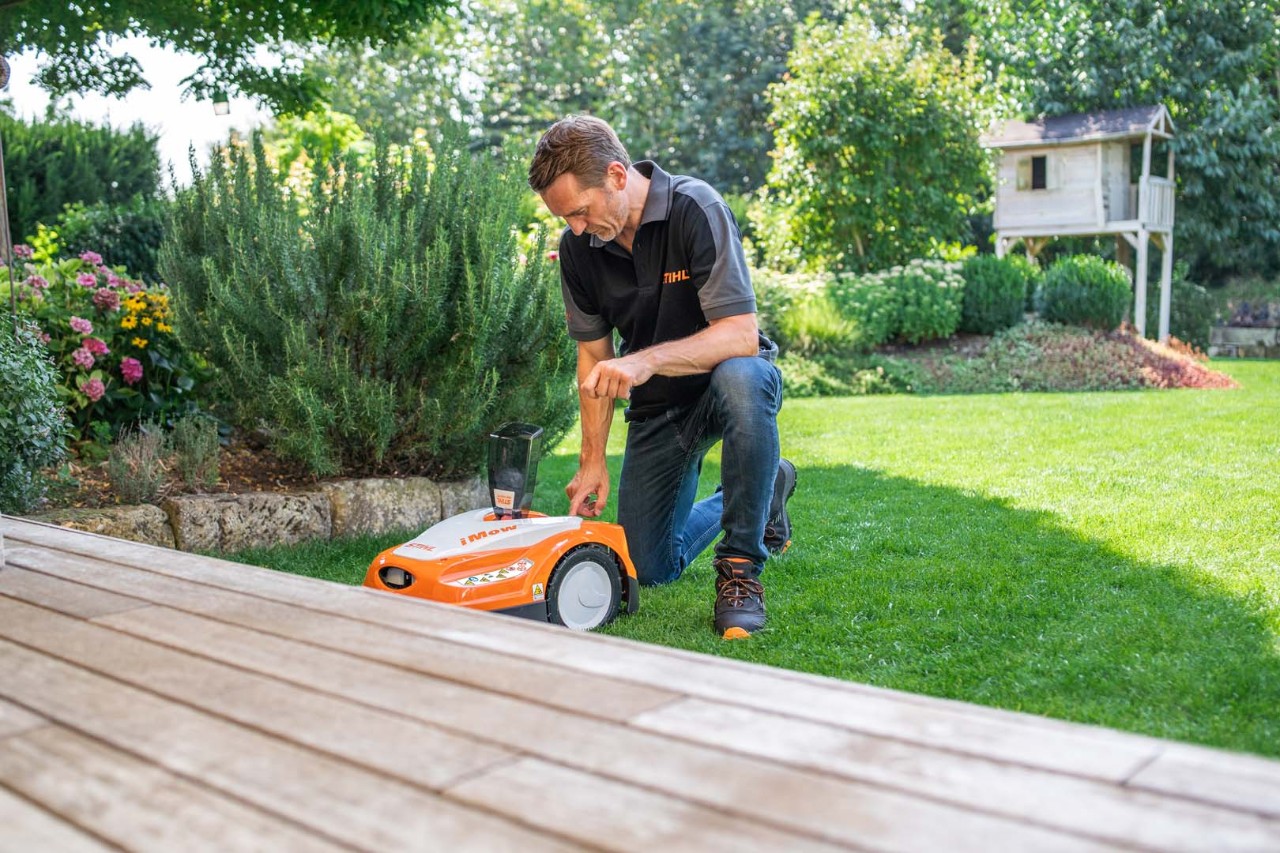 Un homme à genoux sur une pelouse à côté d’un robot de tonte STIHL iMOW®, dans un jardin avec des arbustes et une terrasse