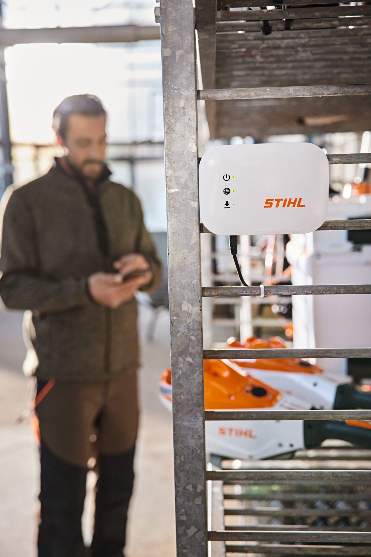 Une STIHL connected Box suspendue à des étagères dans un atelier avec un paysagiste qui interagit avec son smartphone. 
